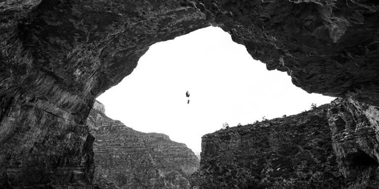 A person is suspended by a rope, rappelling down from a large rocky cliff toward the mouth of a Grand Canyon cave.