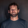 A man with short brown hair and a beard, wearing a dark t-shirt, stands in front of a plain dark background, smiling at the camera. Stephen Eginoire, photographer of Grand Canyon caves